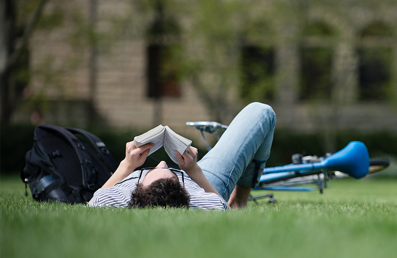 Student reading a book while lying in green grass.
